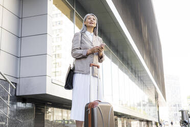 Businesswoman with suitcase holding smart phone while standing outside office building - OIPF01402