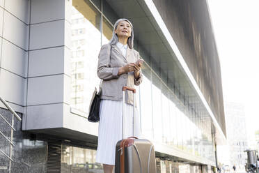 Geschäftsfrau mit Koffer hält Smartphone, während sie vor einem Bürogebäude steht - OIPF01402