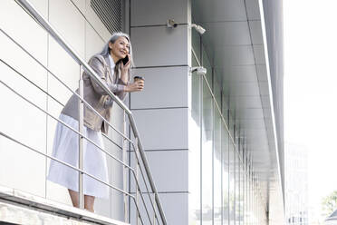 Female professional holding disposable coffee cup while talking on mobile phone at railing - OIPF01407