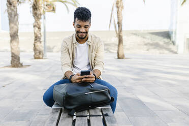 Smiling man with backpack using smart phone on bench - XLGF02346