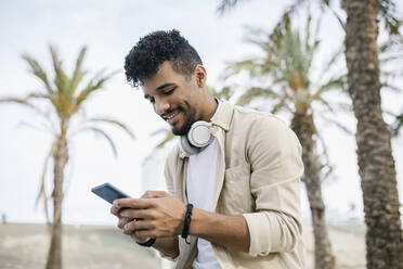 Smiling man with wireless headphones text messaging through mobile phone - XLGF02347