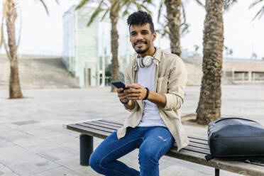 Junger Mann mit Mobiltelefon und Rucksack auf einer Bank sitzend - XLGF02349