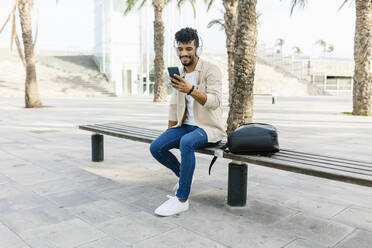 Man listening music through wireless headphones while using mobile phone on bench - XLGF02358