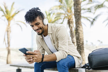 Young man with wireless headphones using mobile phone on bench - XLGF02359