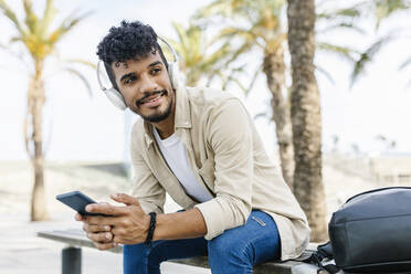 Young man wearing wireless headphones holding mobile phone while sitting on bench - XLGF02360