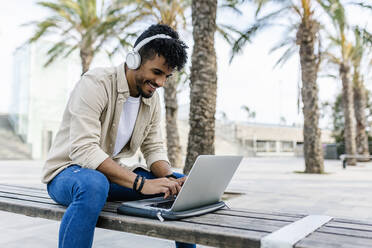 Smiling young man wearing wireless headphones using laptop on bench - XLGF02368