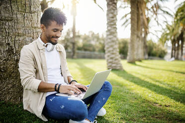 Lächelnder junger Mann, der einen Laptop benutzt, während er an einem Baumstamm sitzt - XLGF02369