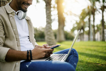 Junger Mann mit Laptop und Smartphone im Park - XLGF02372