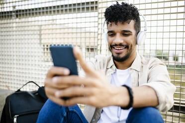 Lächelnder Mann mit drahtlosen Kopfhörern und Mobiltelefon vor einem Zaun - XLGF02375