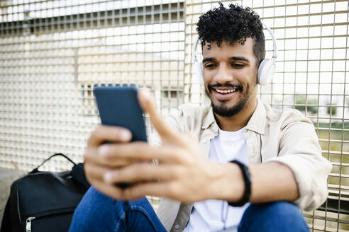 Lächelnder Mann mit drahtlosen Kopfhörern und Mobiltelefon vor einem Zaun - XLGF02375