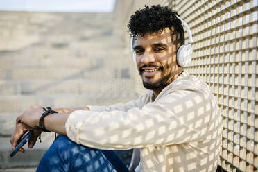 Young man listening music through wireless headphones while sitting by fence - XLGF02377