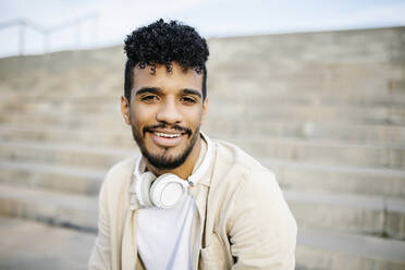 Lächelnder junger Mann mit schwarzem lockigem Haar und drahtlosen Kopfhörern - XLGF02380