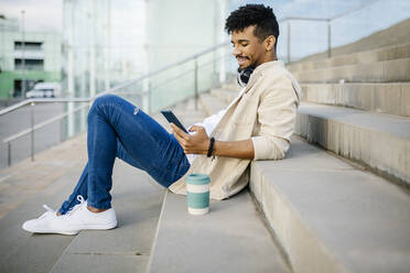 Junger Mann, der ein Mobiltelefon benutzt, während er auf einer Treppe sitzt - XLGF02384