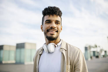Lächelnder junger Mann mit lockigem Haar und drahtlosen Kopfhörern - XLGF02387