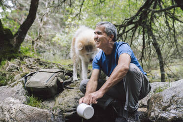 Glücklicher Mann mit Kaffeetasse neben Akita-Hund auf einem Felsen im Wald sitzend - JCCMF04255