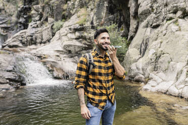Smiling mid adult man talking on smart phone through speaker by pond - JCCMF04270