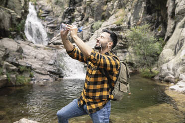 Lächelnder männlicher Tourist, der sich mit einer Flasche am Teich Wasser ins Gesicht schmiert - JCCMF04272