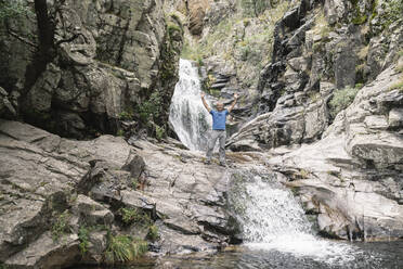 Mature male tourist with arms raised standing on rock - JCCMF04274