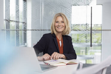 Lächelnde blonde Geschäftsfrau sitzt mit Tagebuch am Schreibtisch - FKF04414