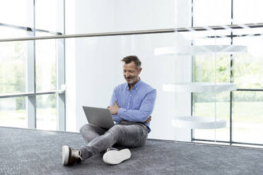 Lächelnde männliche Fachkraft mit Laptop sitzt im Bürokorridor - FKF04424
