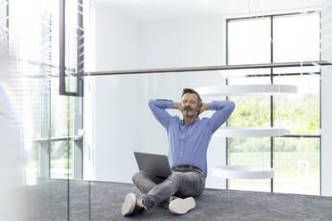 Tired businessman with laptop sitting on ground in corridor - FKF04428