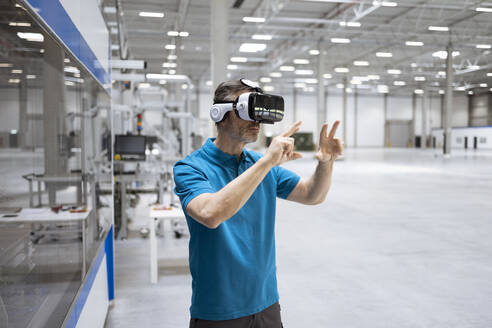 Männlicher Fachmann gestikuliert bei der Verwendung eines Virtual-Reality-Headsets in einer Fabrik - FKF04434