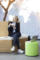Lächelnde weibliche Fachkraft mit Kaffee auf dem Sitz im Büro - FKF04456