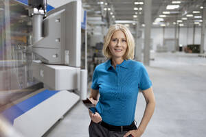 Weibliche Fachkraft lächelt, während sie ein digitales Tablet an einer Maschine hält - FKF04485