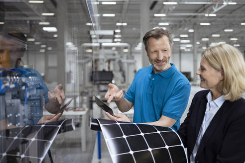 Lächelnde männliche und weibliche Fachleute, die in einer Fabrik über ein Solarpanel diskutieren - FKF04520