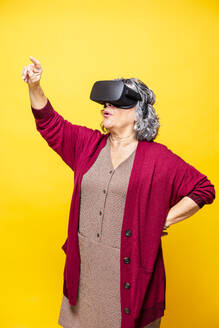 Ältere Frau, die auf etwas zeigt, während sie ein Virtual-Reality-Headset vor gelbem Hintergrund trägt - GPF00084