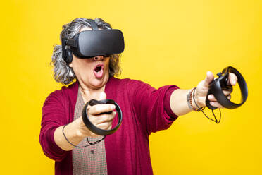 Frau hält Joystick, während sie ein Virtual-Reality-Headset vor gelbem Hintergrund trägt - GPF00086