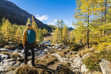 Älterer männlicher Wanderer mit Rucksack, der die Berge im Herbst im Val Masino, Sondrio, Italien, betrachtet - MCVF00911