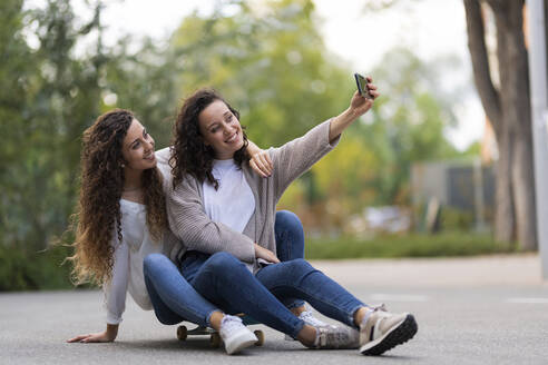 Lächelnde Frau, die ein Selfie mit einer Freundin durch ein Smartphone macht, während sie auf einem Skateboard sitzt - JPF00442