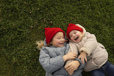 Mädchen mit Strickmütze kitzelt ihre Schwester, während sie auf dem Spielplatz im Gras liegt - SSGF00072
