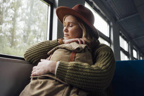 Mittlere erwachsene Frau mit geschlossenen Augen, die einen Rucksack hält, während sie im Zug reist - VPIF05116