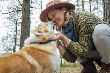 Dog licking face of woman crouching in forest - VPIF05147