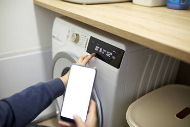 Woman operating washing machine through smart phone in utility room at home - ZEDF04285