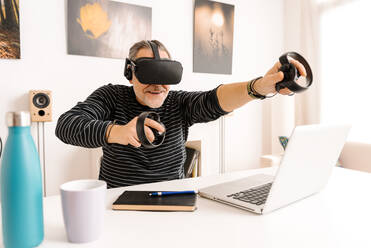 Glücklicher reifer Mann, der einen Virtual-Reality-Simulator trägt und eine Fernbedienung hält, während er am Tisch sitzt - GPF00096