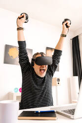 Cheerful man with arms raised wearing virtual reality simulator at home - GPF00100