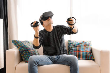 Mature man wearing virtual reality simulator holding remote control while sitting on sofa - GPF00106