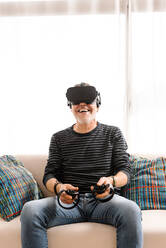 Happy mature man playing with virtual reality simulator on sofa at home - GPF00107