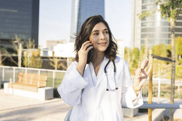 Lächelnde Ärztin, die mit einem Smartphone vor einem Gebäude spricht - JCCMF04275