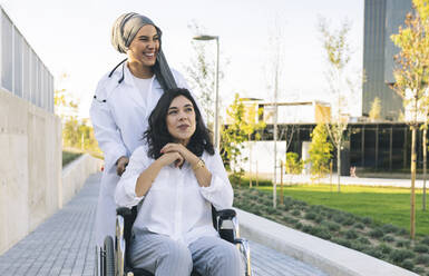 Cheerful female healthcare worker standing behind disabled woman sitting on wheelchair - JCCMF04282