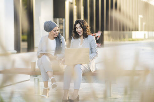 Cheerful female professionals discussing over laptop while sitting on bench - JCCMF04289