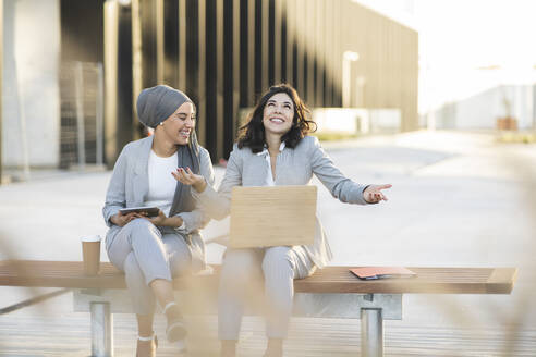 Happy businesswoman gesturing while sitting by female colleague on bench - JCCMF04290