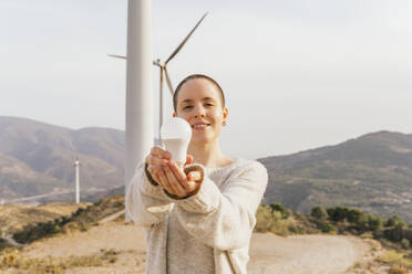 Lächelnde Frau, die eine Glühbirne hält, während sie an einem sonnigen Tag in einem Windpark steht - MGRF00545