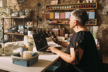 Ältere Künstlerin mit Laptop und Mobiltelefon in der Werkstatt - GPF00126