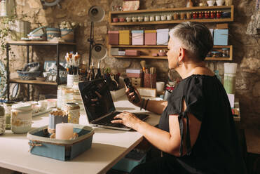 Mature female artist with laptop using mobile phone in workshop - GPF00126