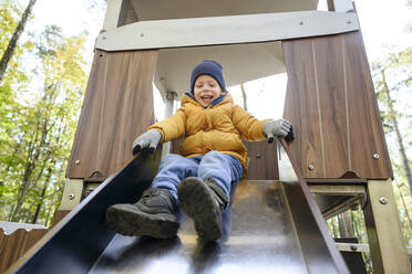Lächelnder Junge in warmer Kleidung rutscht auf einer Rutsche im Park - EYAF01789