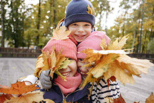 Lächelnde Geschwister spielen mit trockenem Blatt im Herbst - EYAF01800